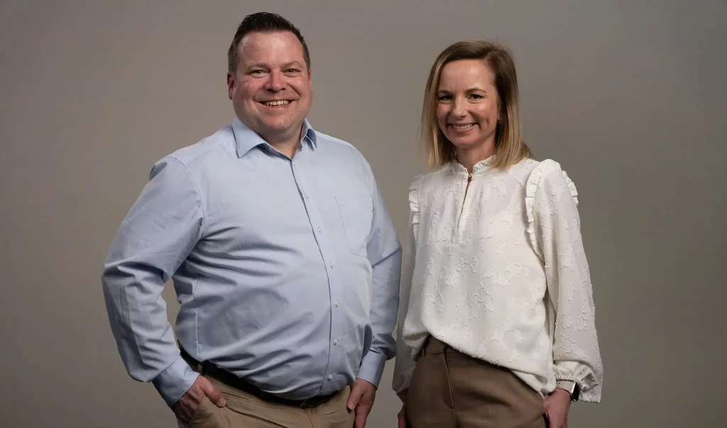 Angela Simonis und Christian Kempf stehen vor einem grauen Hintergrund und lächeln in die Kamera